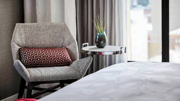 Ett hotellrum med en grå fåtölj, en mönstrad kudde, ett litet bord med en vas och en säng med ett mönstrat överkast.