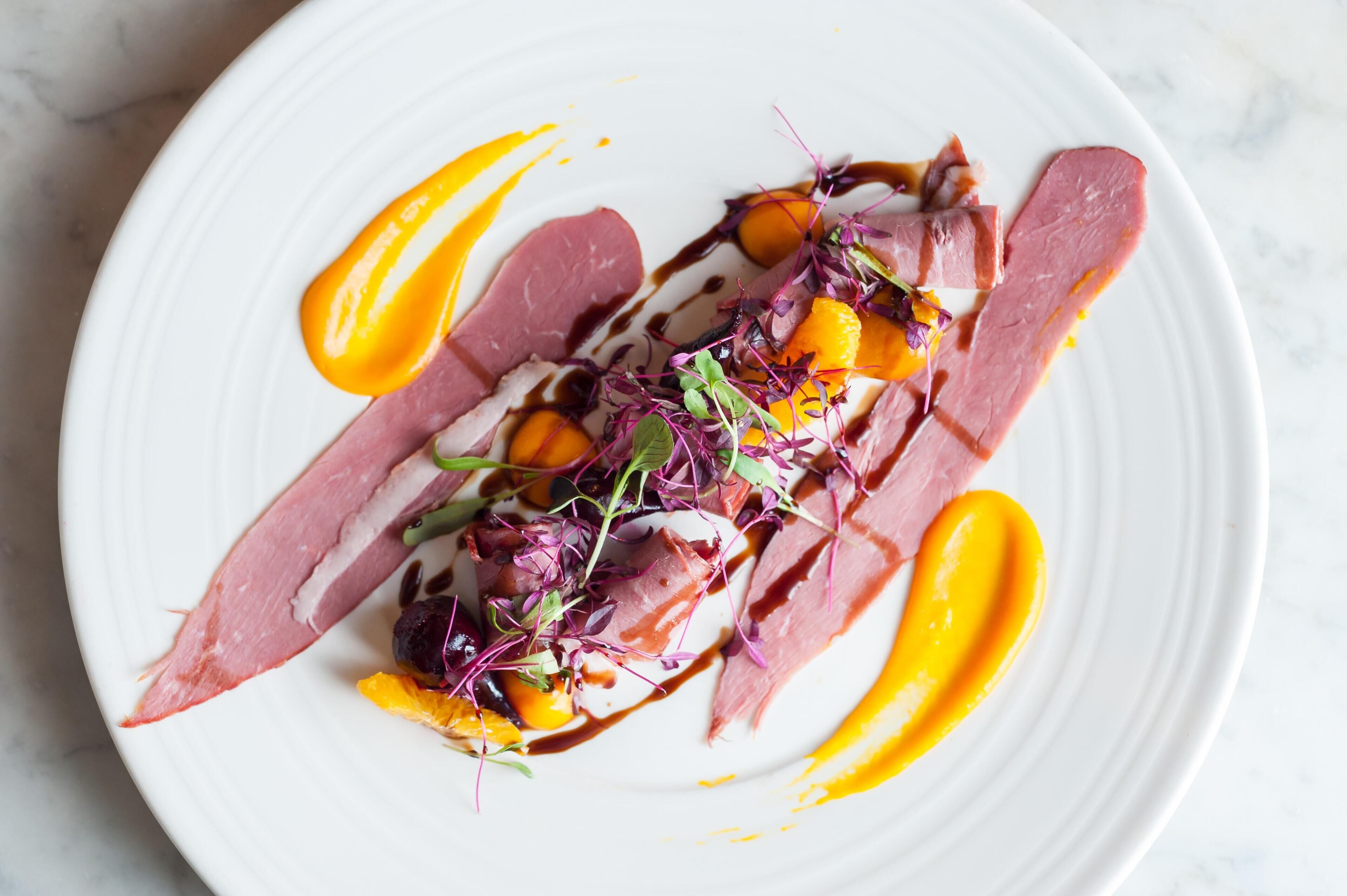 A white plate with a dish of food, including slices of meat and vegetables, and a drizzle of sauce.