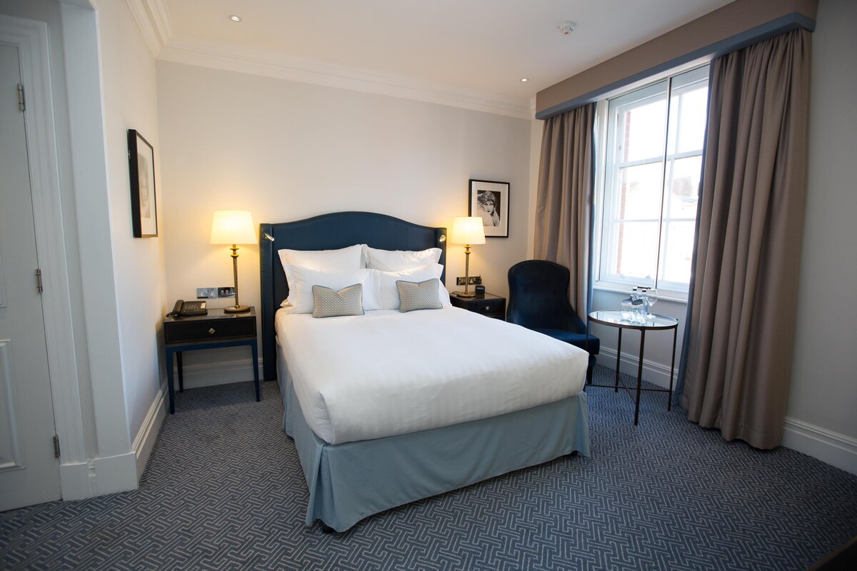 A hotel room with a large bed, two bedside tables with lamps, a chair, a small table, and a window with curtains.