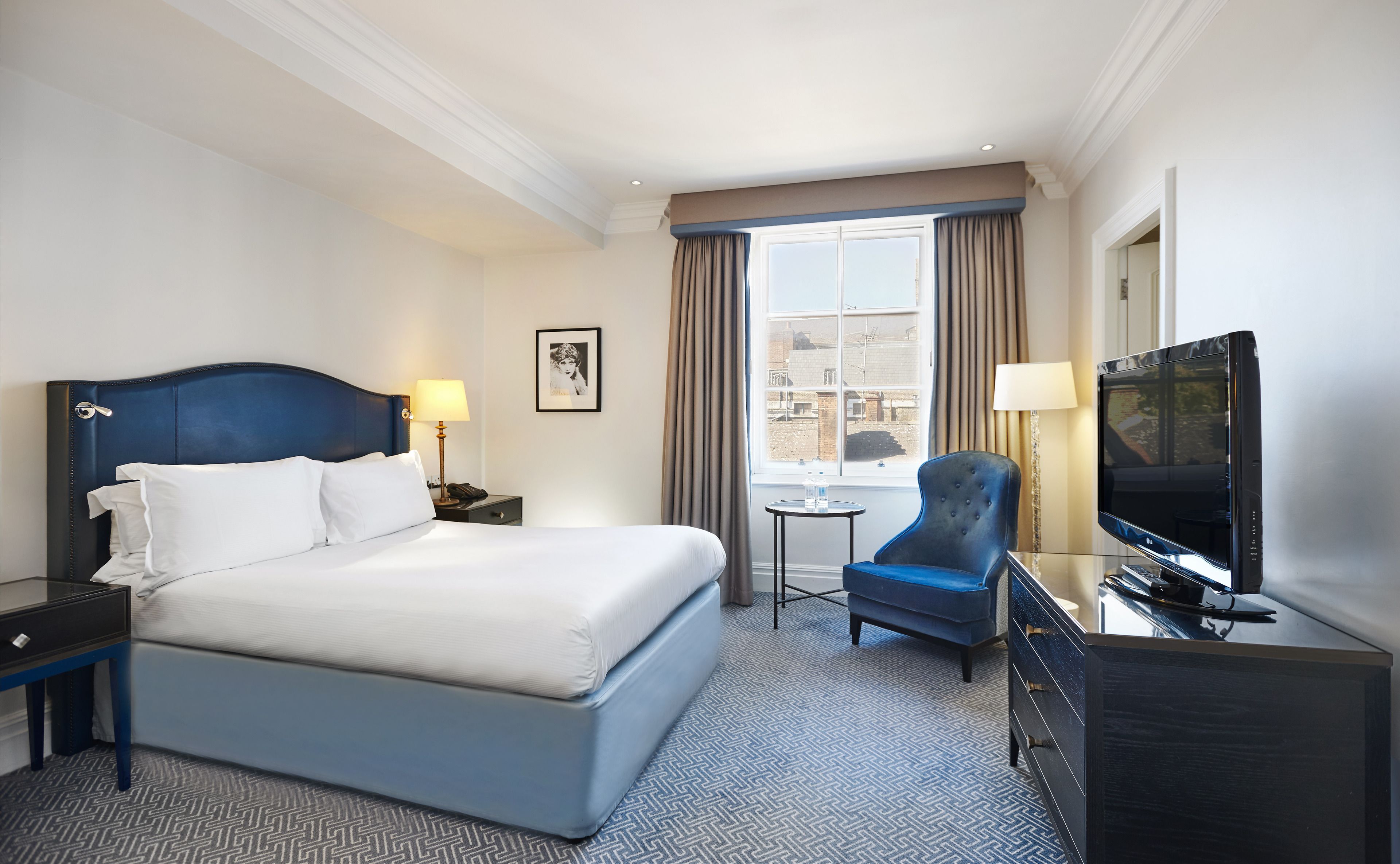 A hotel room with a large bed, a desk with a TV, a chair, and a small table with a lamp.