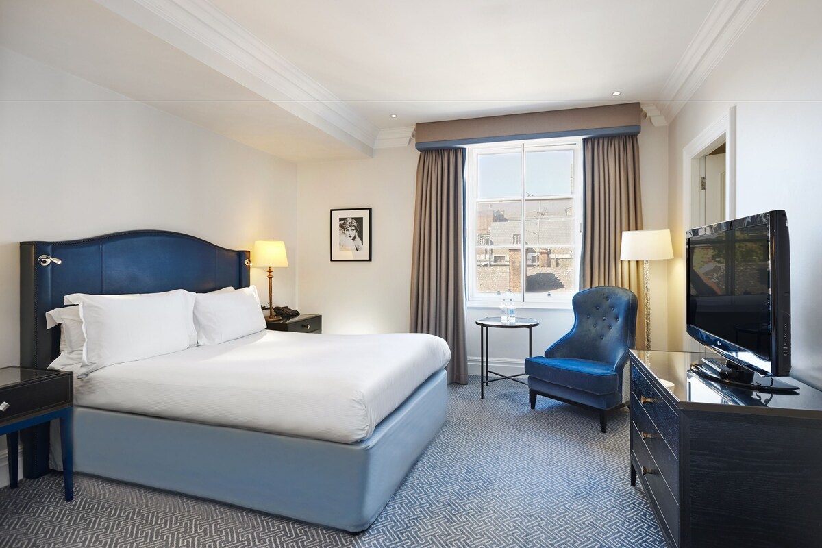 A hotel room with a large bed, a desk with a TV, a chair, and a small table with a lamp.