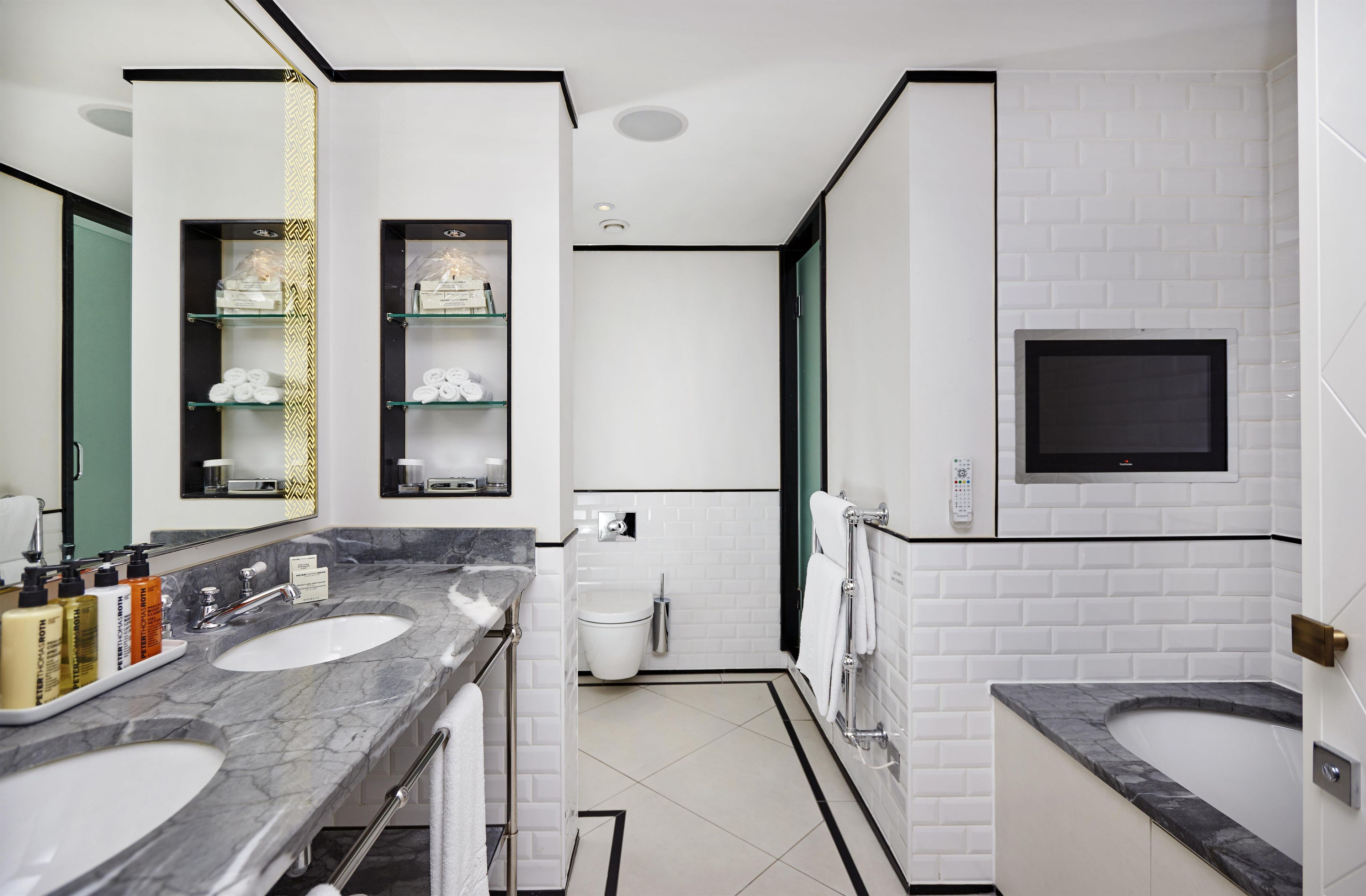 A modern bathroom with two sinks, a toilet, a shower, and a TV mounted on the wall.