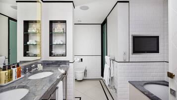 A modern bathroom with two sinks, a toilet, a shower, and a TV mounted on the wall.