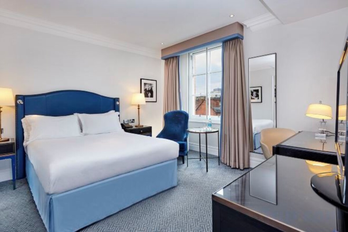 A hotel room with a large bed, a desk, a chair, a small table, and a window with curtains.