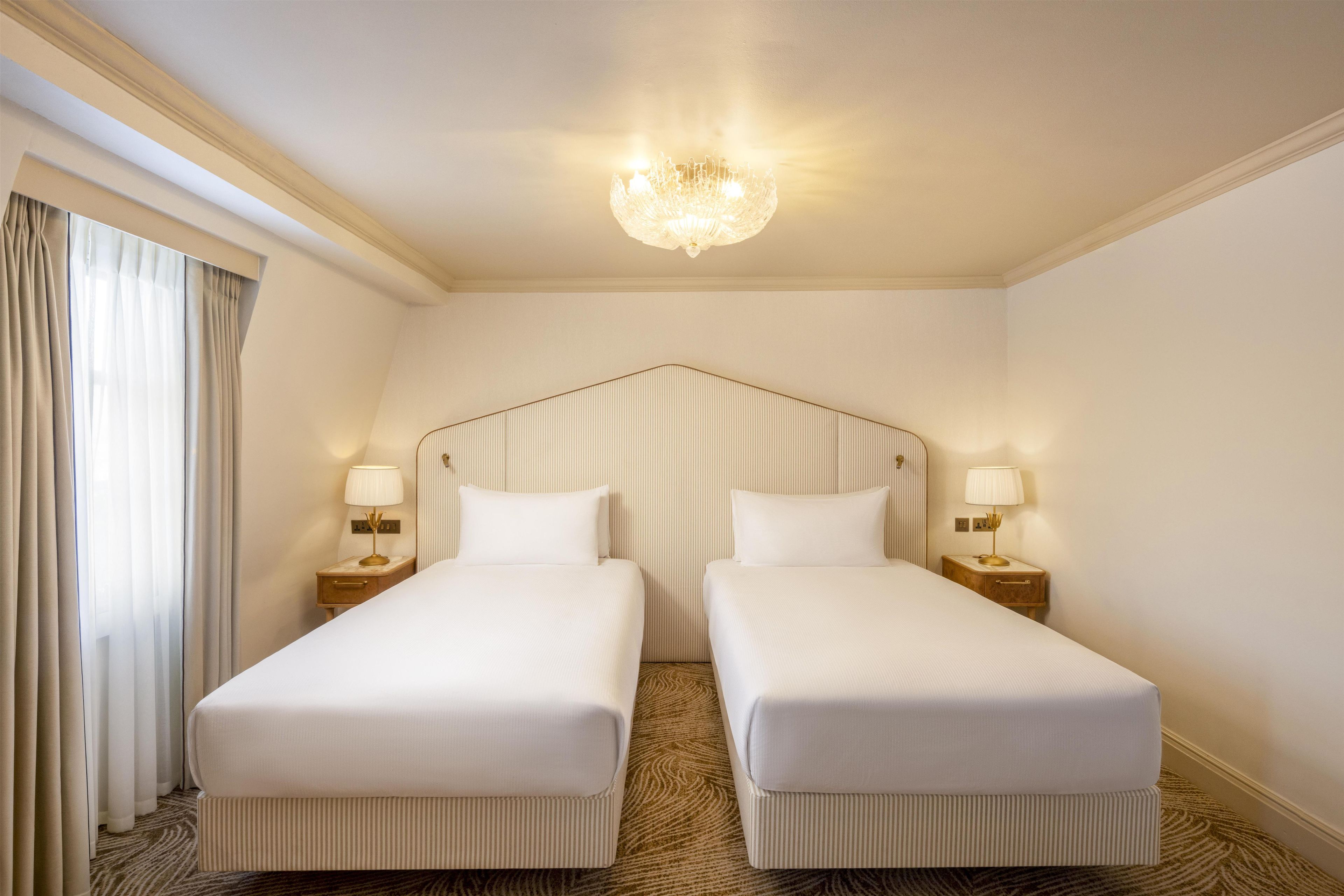 A hotel room with two single beds, a chandelier, and a window with curtains.