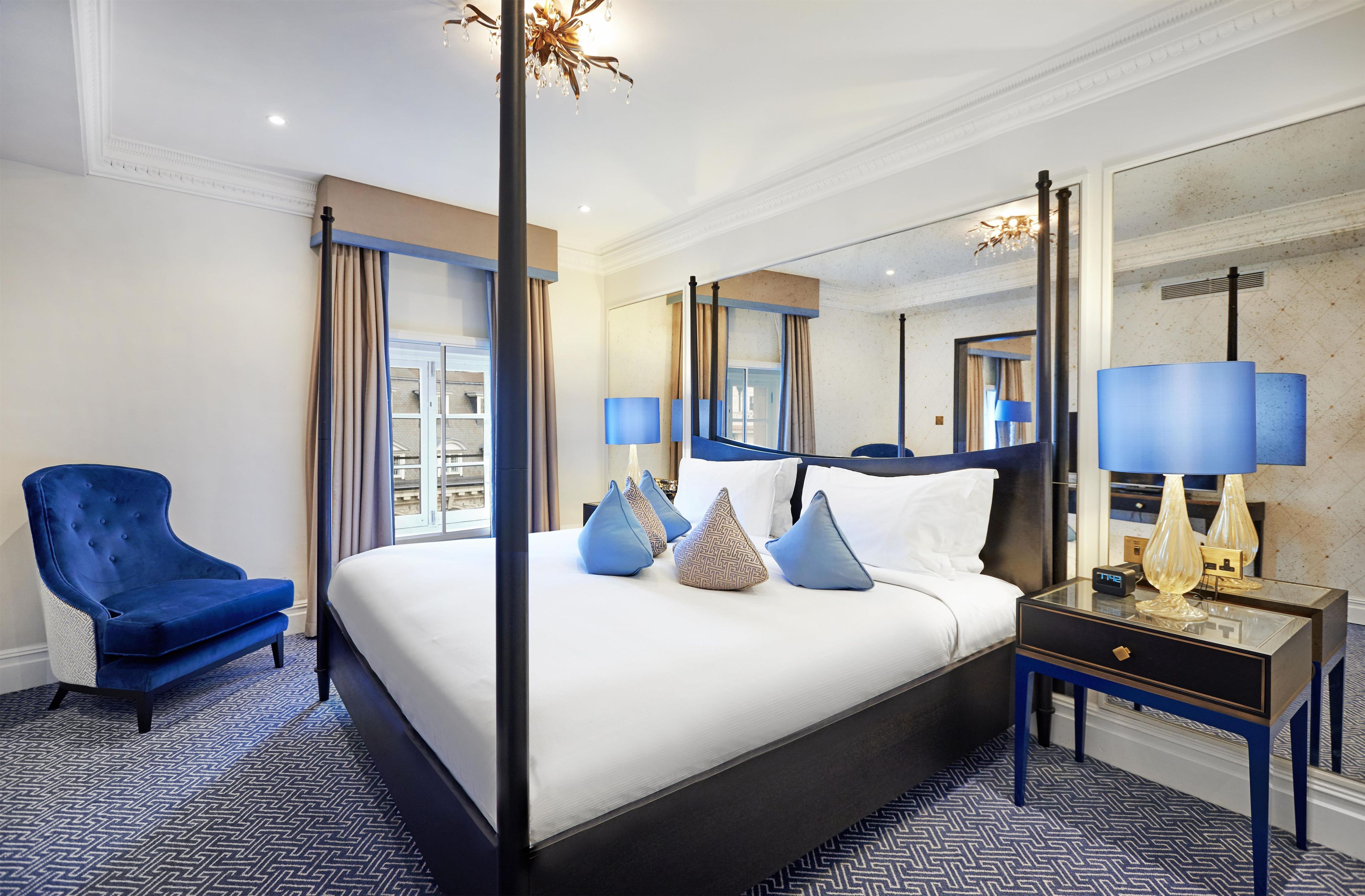 A hotel room with a large bed, a blue armchair, a nightstand with lamps, and a mirror.