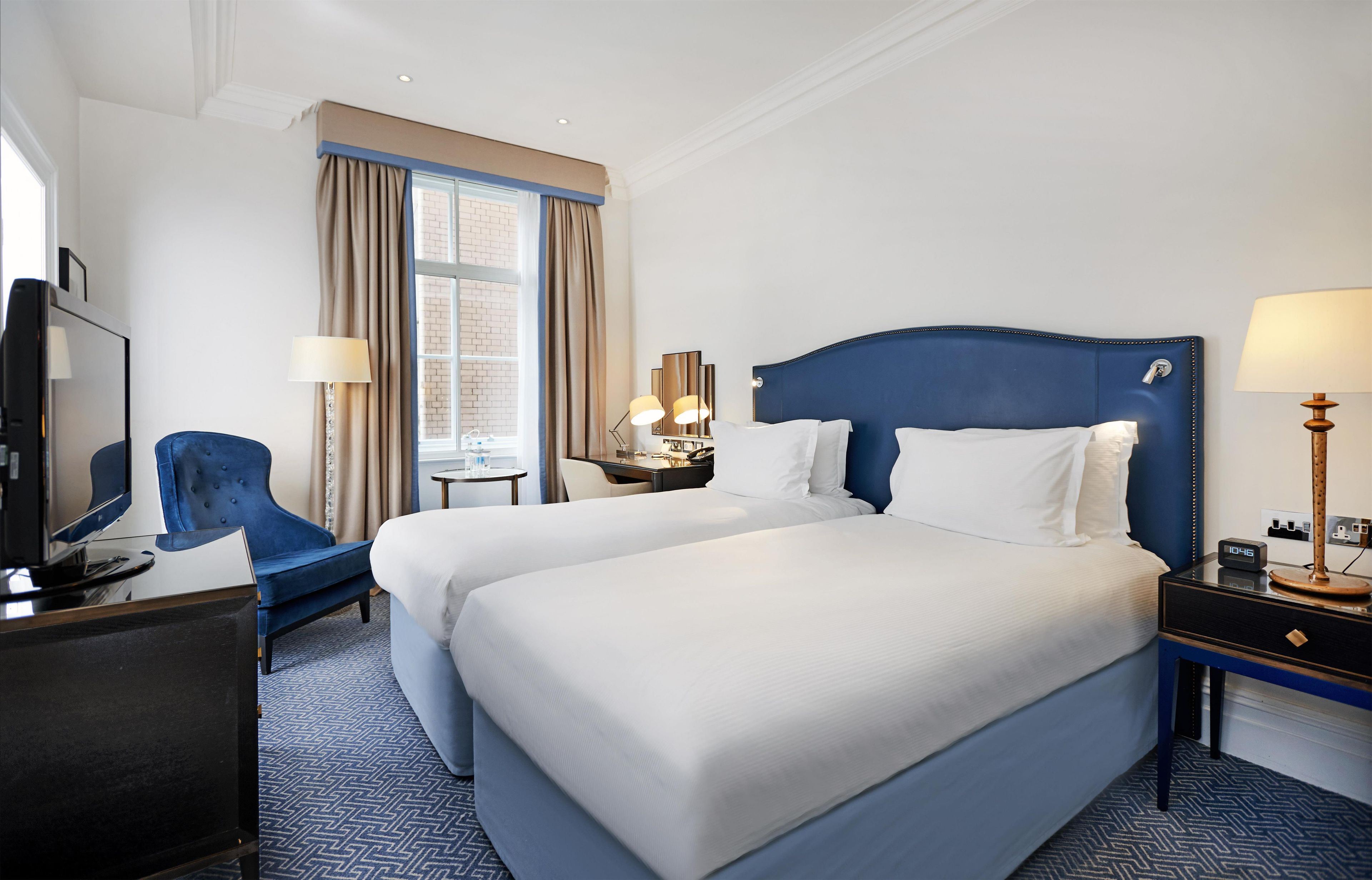 A hotel room with two beds, a desk, a chair, a TV, and a window with curtains.