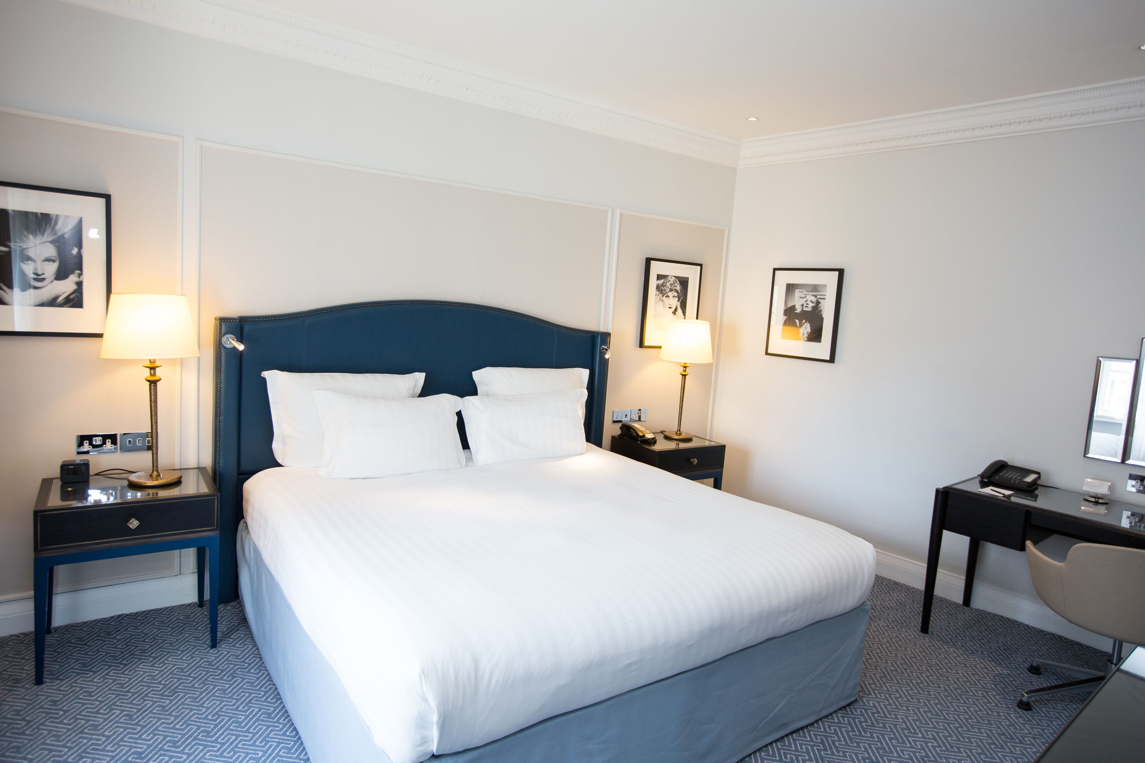 A hotel room with a large bed, two bedside tables with lamps, a desk with a chair, and framed pictures on the walls.