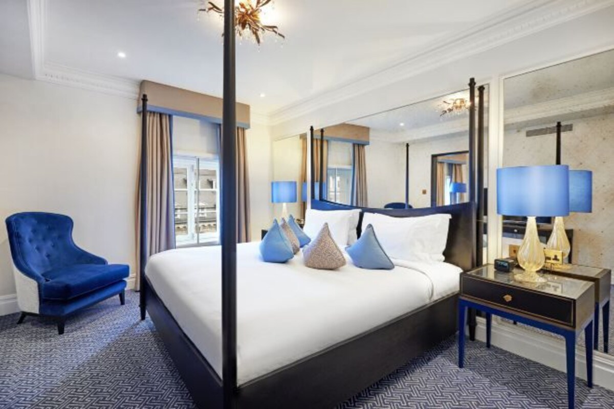 A hotel room with a large bed, a blue armchair, a nightstand with lamps, and a large mirror.