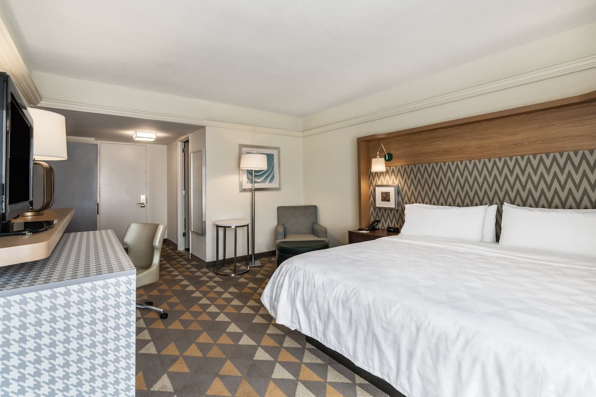 A hotel room with a large bed, a desk with a lamp, a chair, a small round table, and a television.