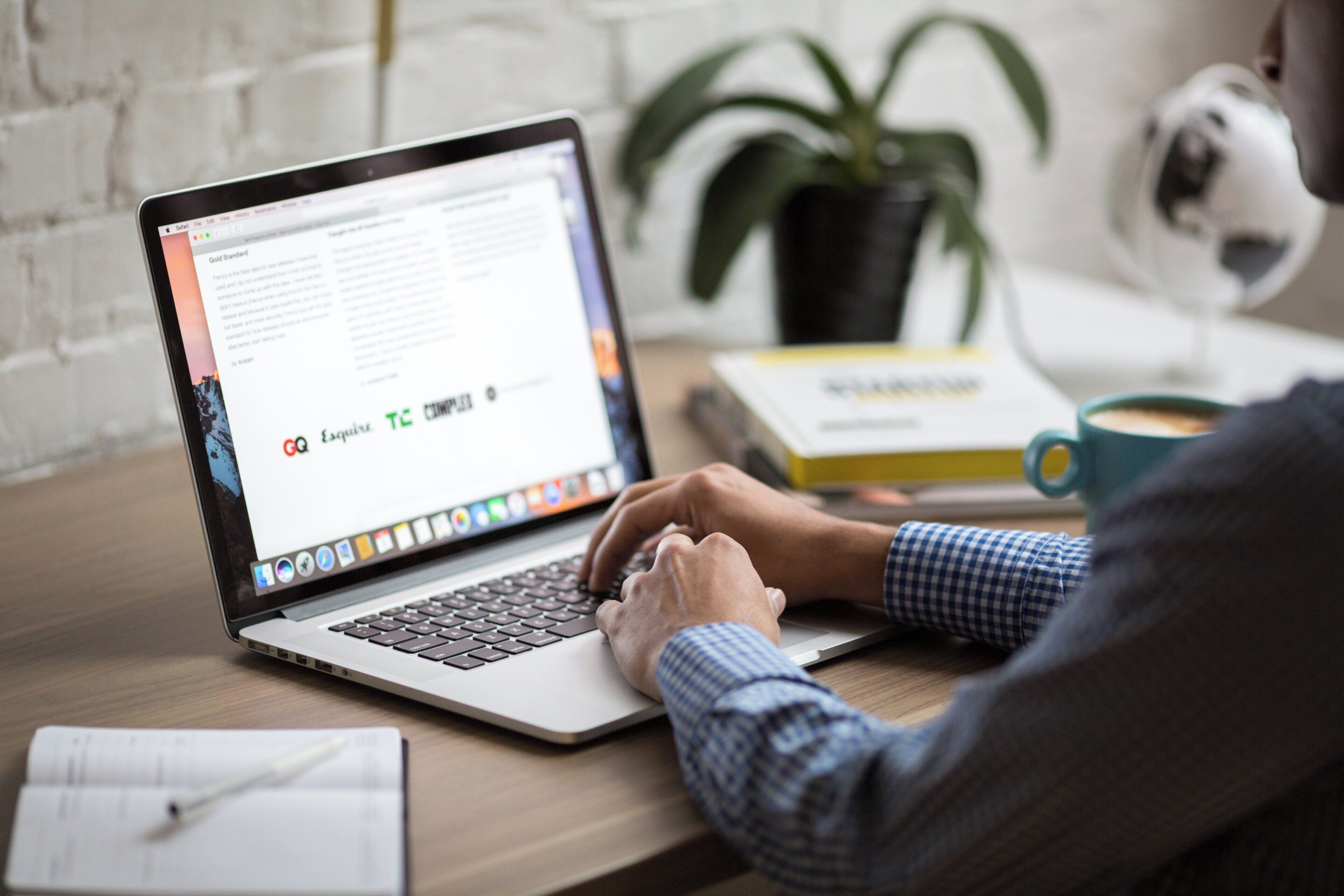 A person using a laptop on a wooden desk with a notebook, a mug, and a plant in the background.