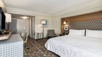 A hotel room with a bed, a desk with a television, a chair, a small table, and a wall-mounted lamp.