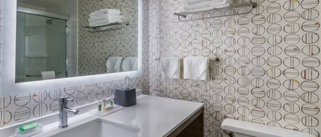 A bathroom with a white sink, mirror, and toilet. There are towels on a shelf and a rack, and toiletries on the sink.