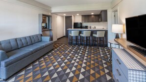 A modern hotel room with a sofa, a TV, a kitchen area with bar stools, and a patterned floor. - Holiday Inn & Suites Pittsfield-Berkshires by IHG (Pittsfield)