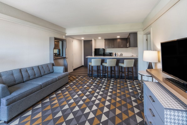 Une chambre d’hôtel moderne dotée d’un canapé, d’une télévision, d’un coin cuisine avec des tabourets de bar et d’un sol à motifs.