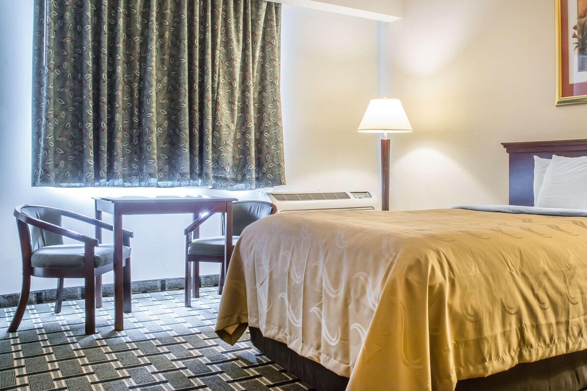 A hotel room with a bed, a desk, two chairs, a lamp, and a window with curtains.