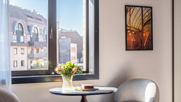 Habitación de hotel moderna con un ventanal grande, una mesa redonda con un florero, dos sillones grises, una cama y una obra de arte enmarcada en la pared.
