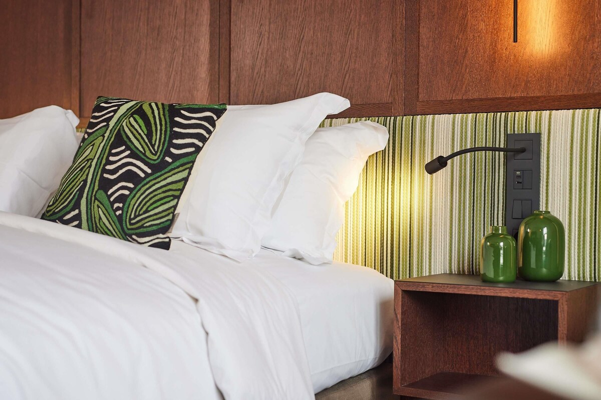 A bed with white linens, a green patterned pillow, and a bedside table with two green vases.