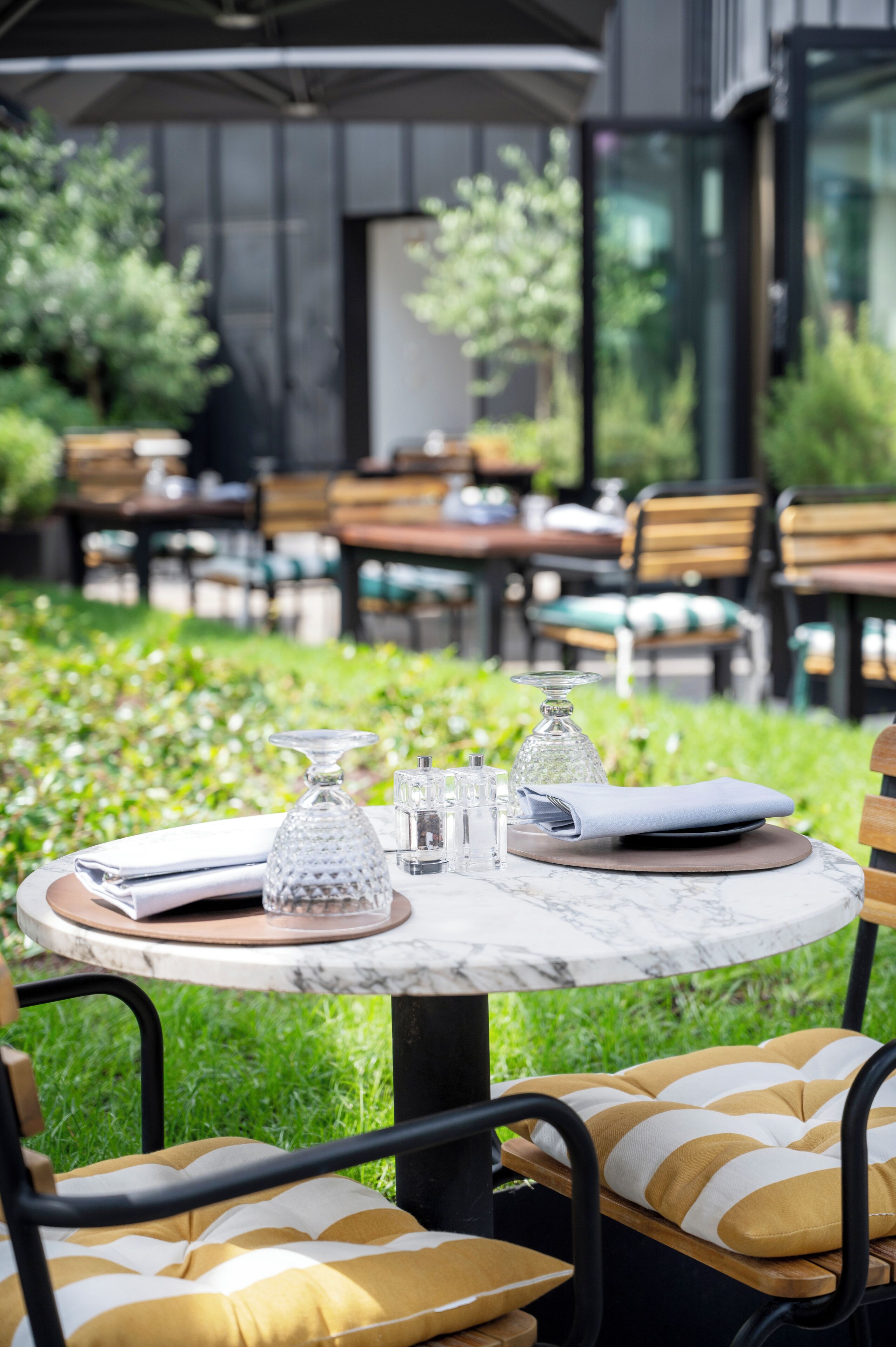 Un área de comedor al aire libre con mesas redondas, sillas y una mesa con cubierta de vidrio, servida con servilleta, cristalería y un menú.