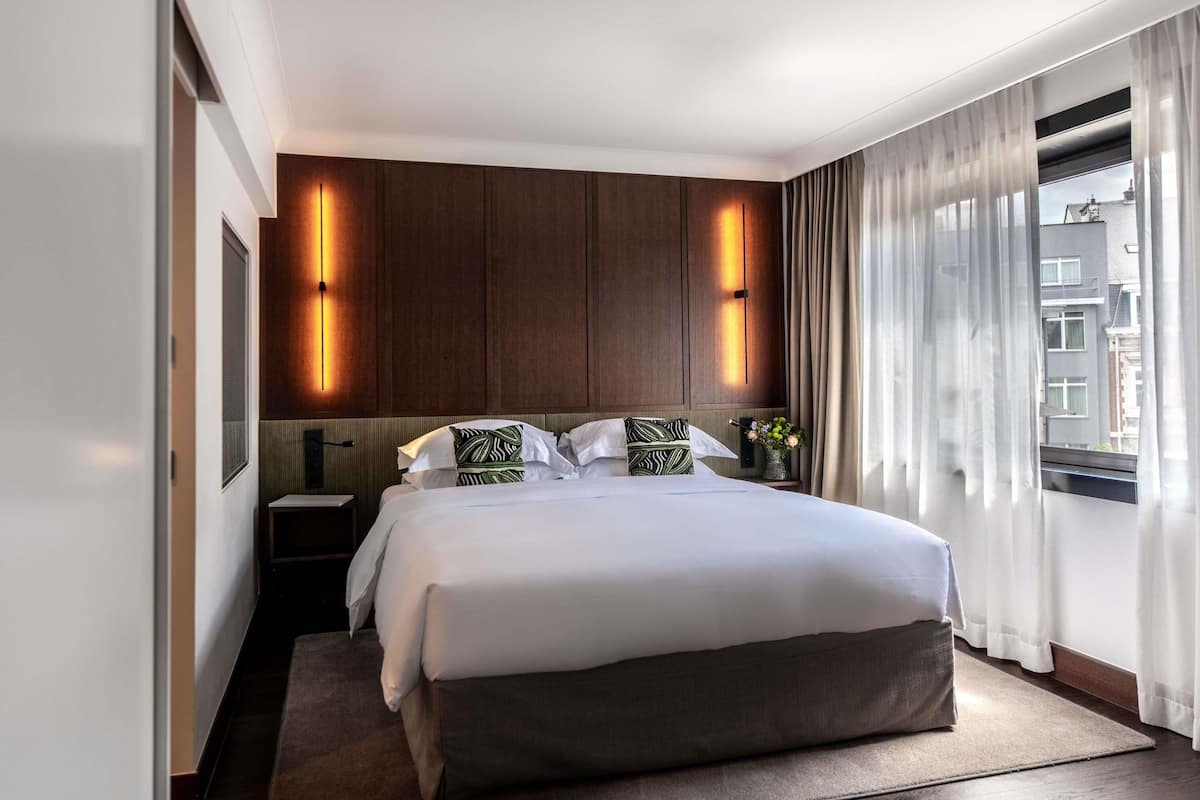 A modern hotel room with a large bed, wooden paneling on the walls, and a view of buildings through the window.