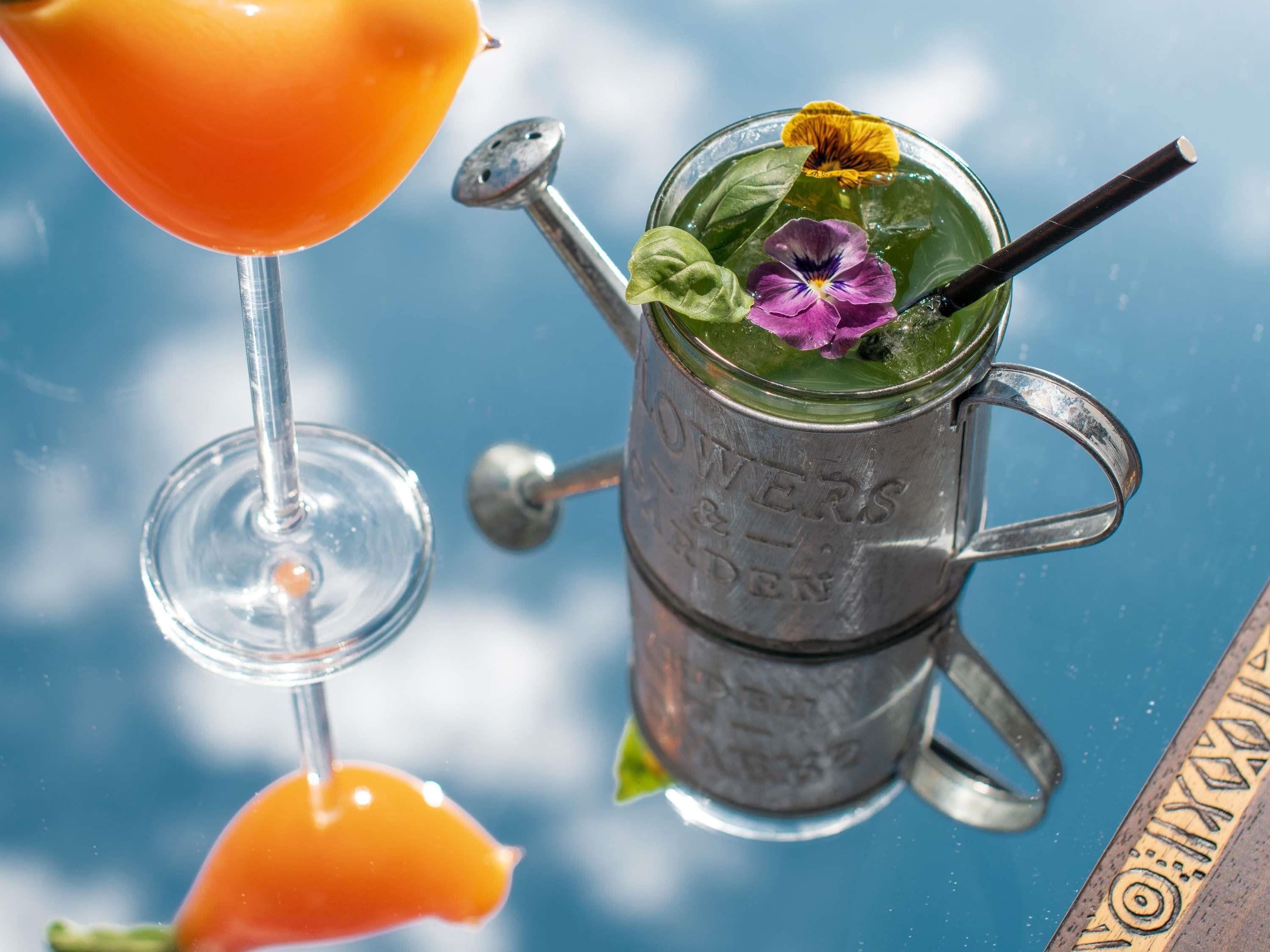 A glass of orange liquid with a straw, a metal pitcher with a handle, and a flower on top.