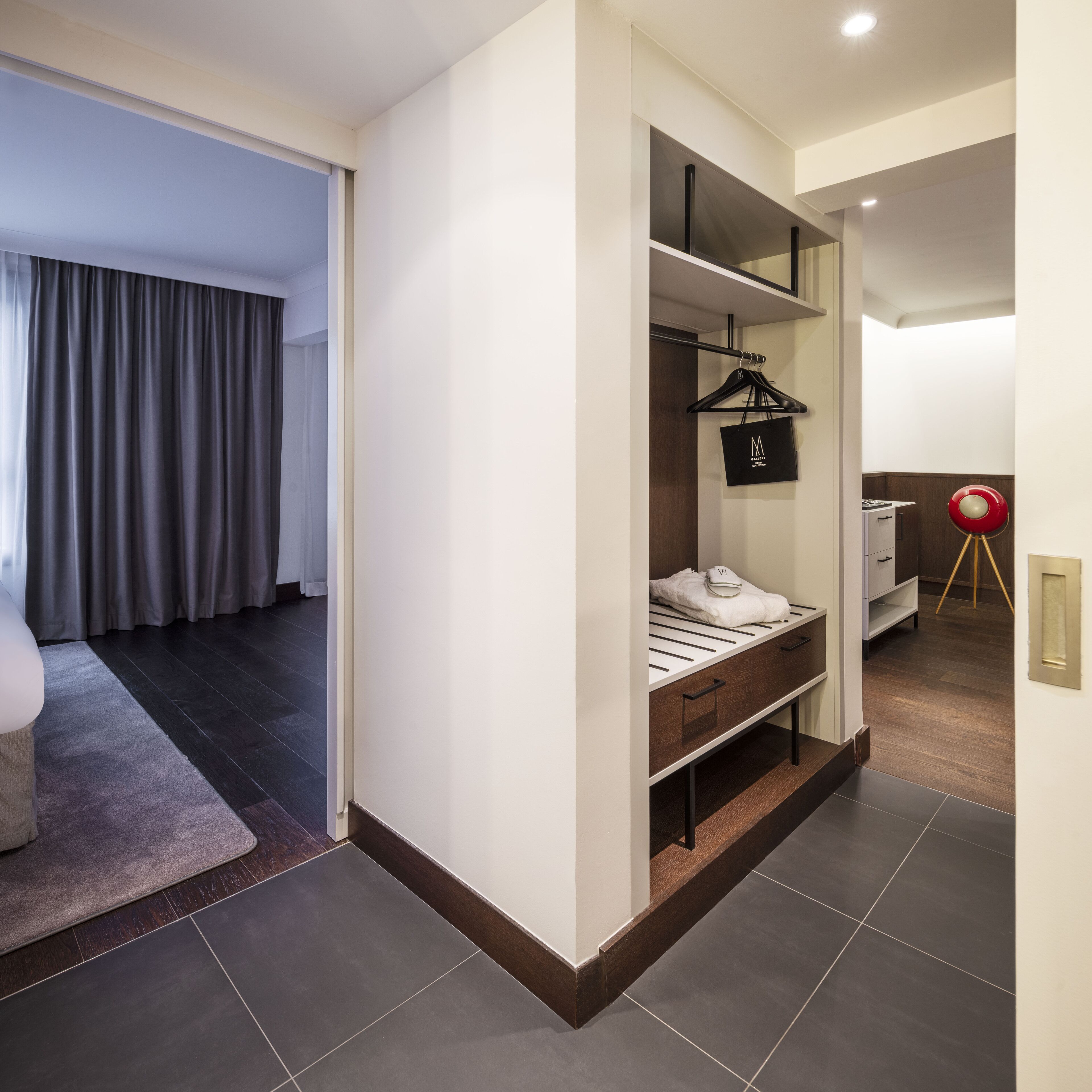Une chambre d’hôtel moderne dotée d’un dressing, d’un lit avec une tête de lit blanche et d’une lampe rouge posée sur un pied de lampe.