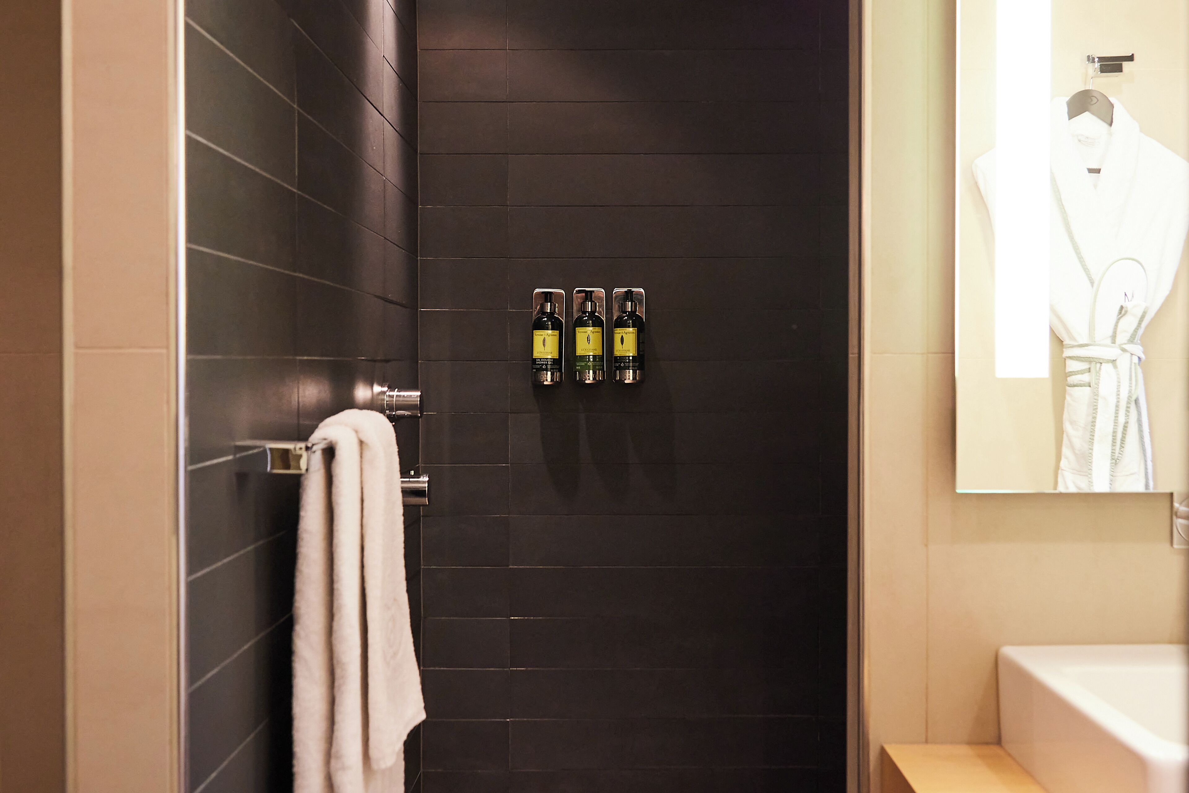 A bathroom with dark tiled walls, a white towel hanging on a rack, and three bottles of hand sanitizer on the wall.