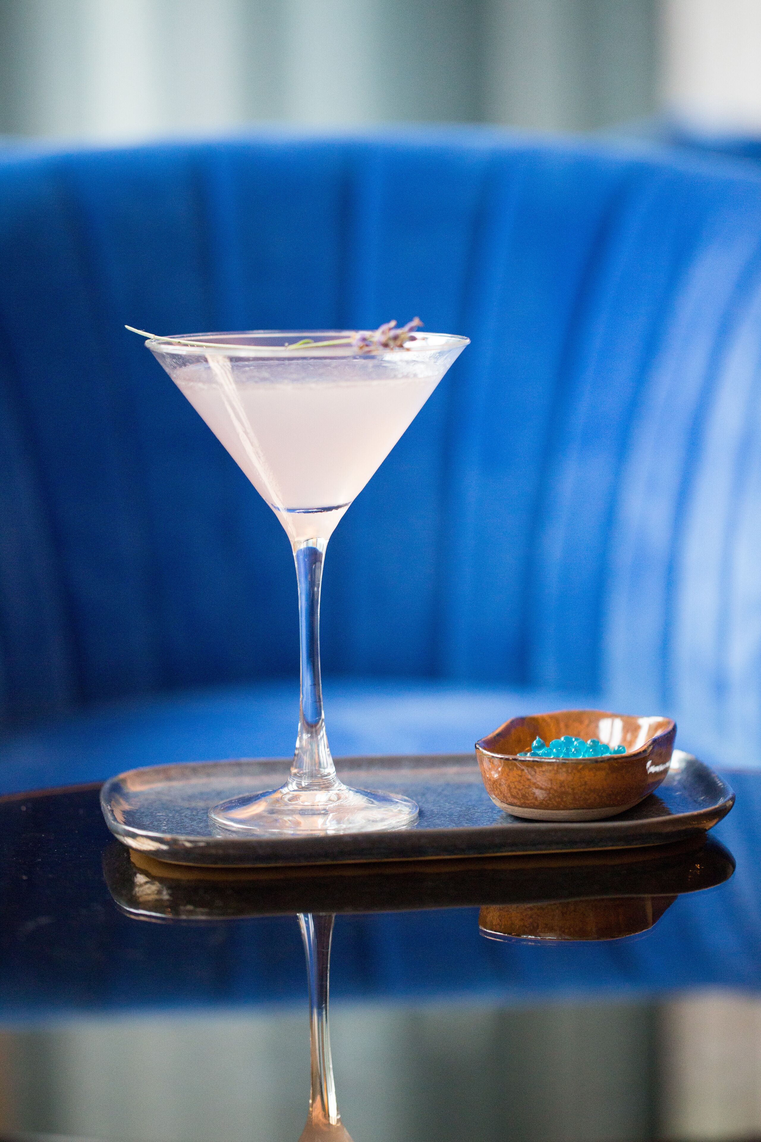 Un verre à martini posé sur un plateau réfléchissant, avec un petit bol de perles bleues à côté, le tout sur un canapé bleu en arrière-plan.