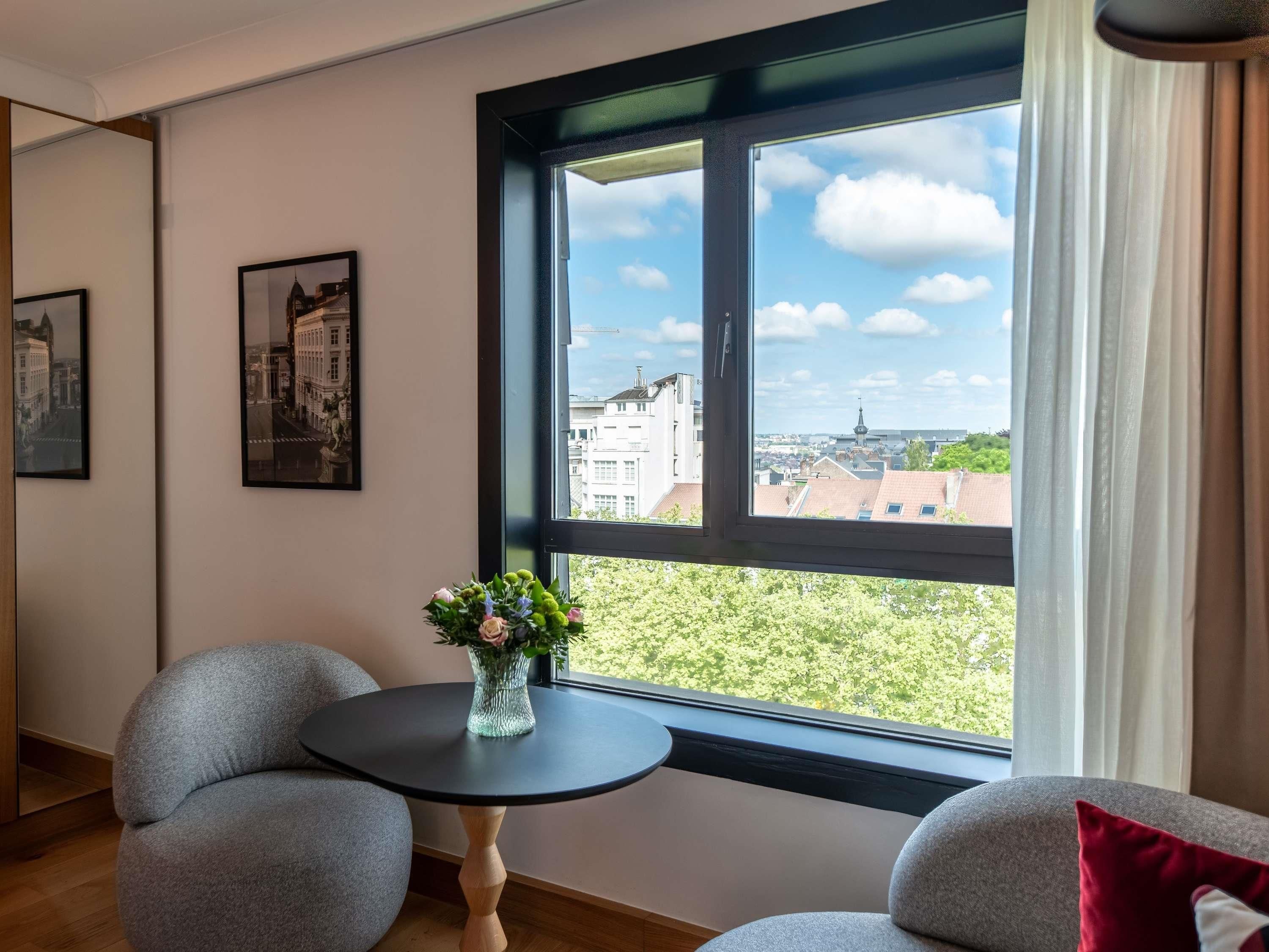 Una habitación con un gran ventanal, dos sillas grises, una pequeña mesa redonda con un jarrón de flores y dos cuadros en la pared.