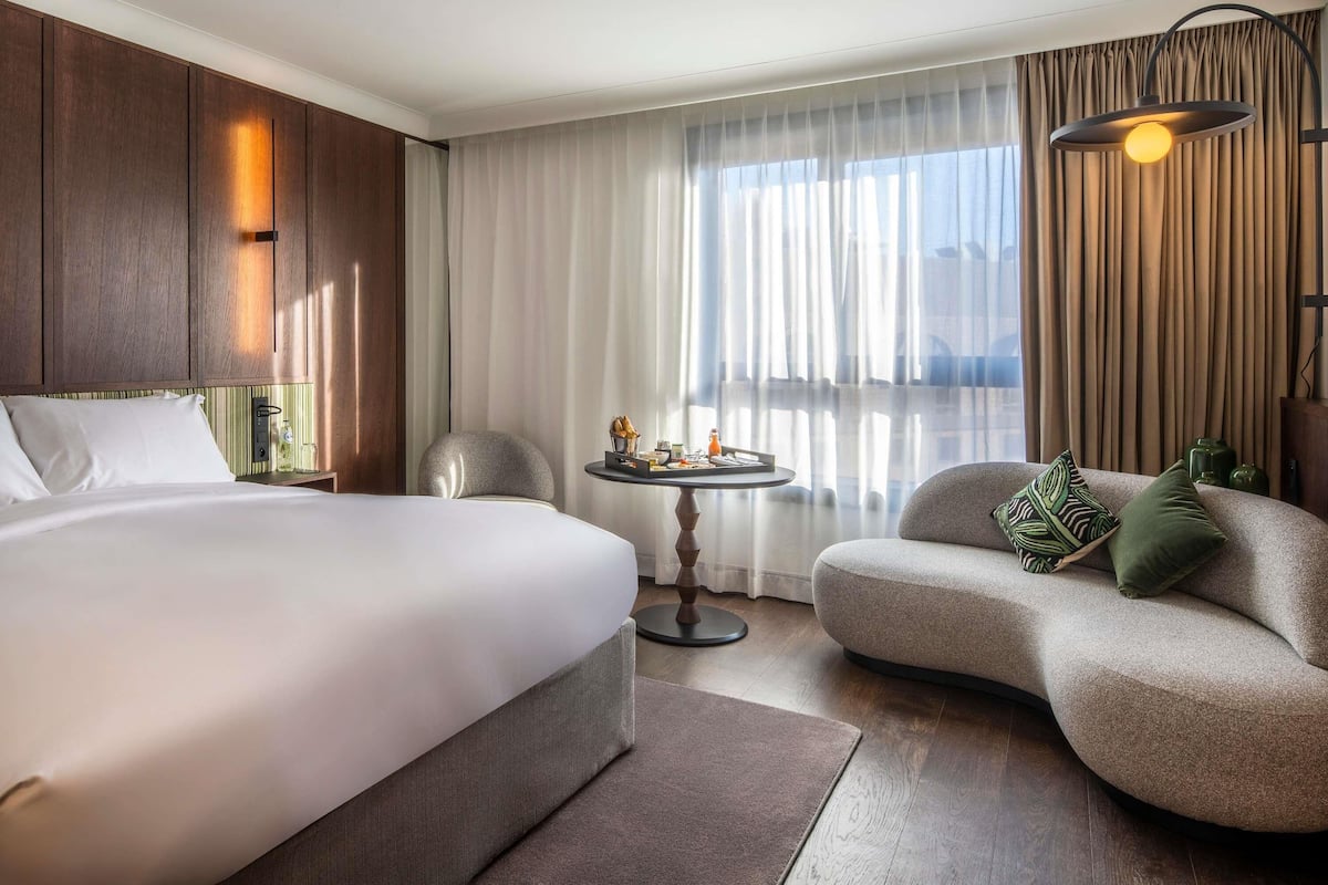 Une chambre d’hôtel avec un grand lit, un canapé, une petite table ronde avec des éléments pour le petit-déjeuner, une lampe et une vue sur la ville à travers des rideaux légers.