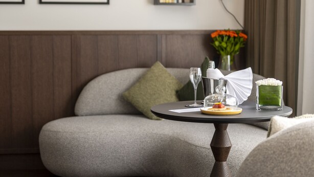 A cozy seating area with a cushioned bench, a small round table with a glass dome, and a wall-mounted lamp. - Le Louise Hotel Brussels - MGallery (Brussels)