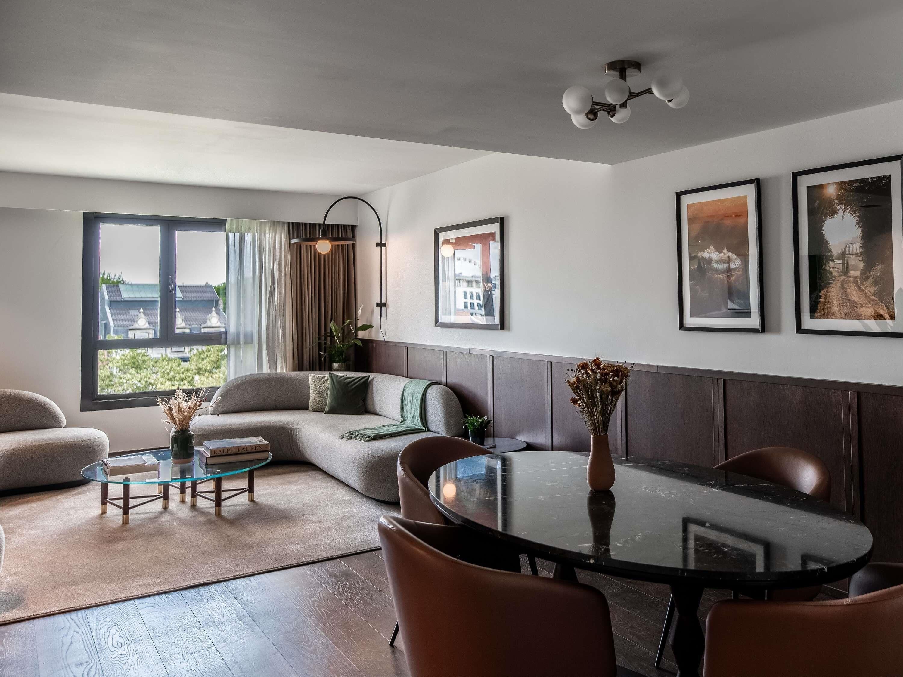 Un salon moderne comprenant un canapé, une table basse en verre, une table à manger avec des chaises, une grande fenêtre avec des rideaux et des tableaux encadrés accrochés au mur.