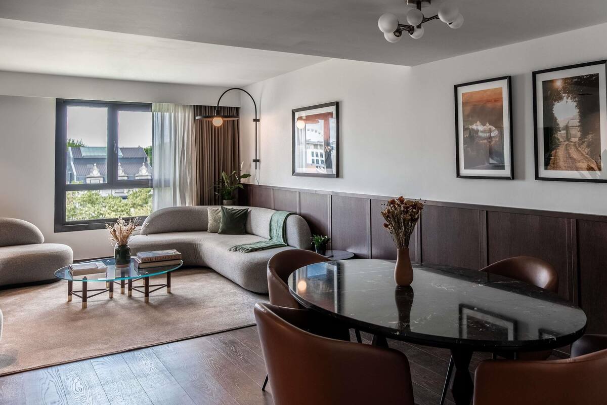 A modern living room with a sofa, a glass coffee table, a dining table with chairs, a large window with curtains, and framed pictures on the wall.