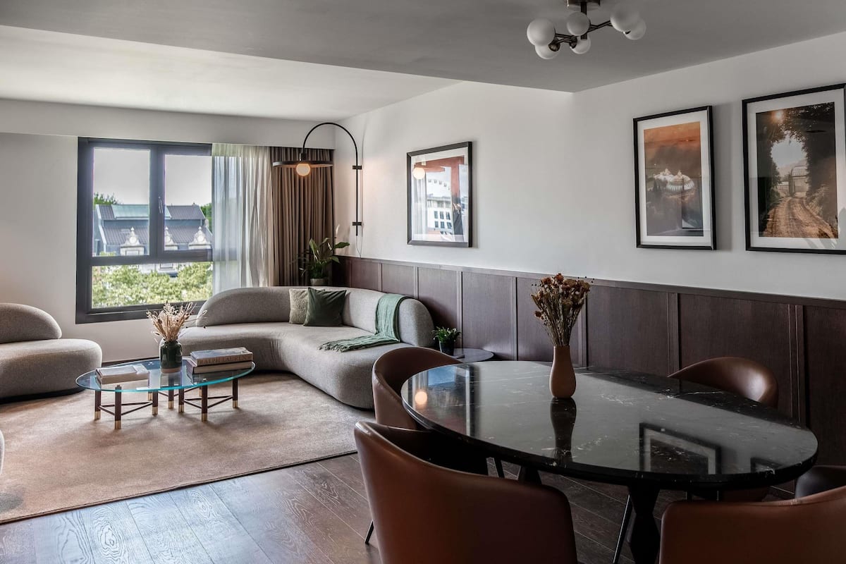 Ein modernes Wohnzimmer mit einer Couch, einem Glastisch, einem Esstisch mit Stühlen, einem großen Fenster mit Vorhängen und gerahmten Bildern an der Wand.