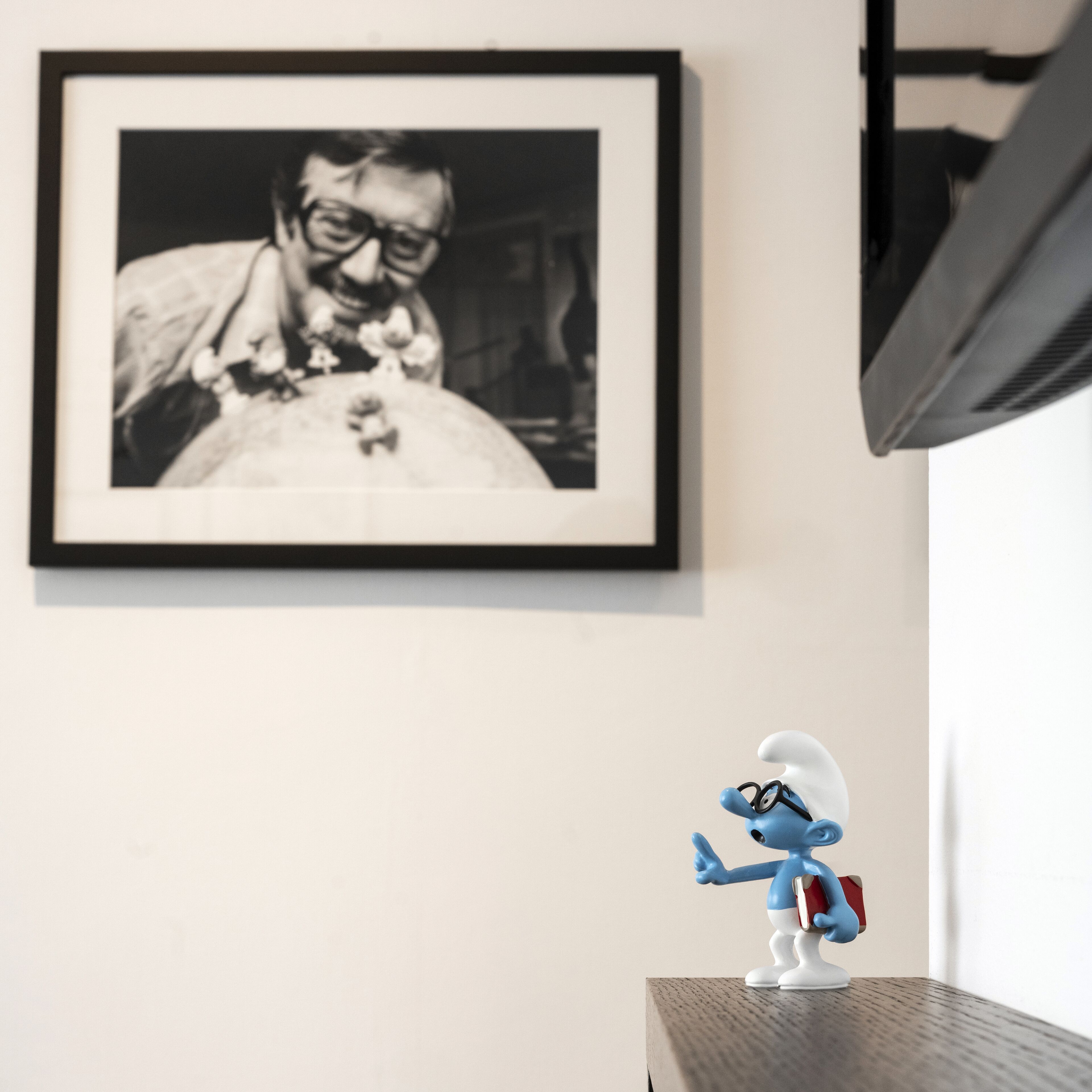 Uma fotografia a preto e branco emoldurada numa parede, uma pequena estatueta sobre uma mesa, e uma janela com persianas.