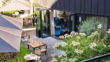 An outdoor patio with tables and chairs, a glass door entrance, and a garden with white flowers.