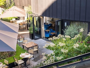 Un patio al aire libre con mesas y sillas, un acceso por puerta de vidrio y un jardín con flores blancas.