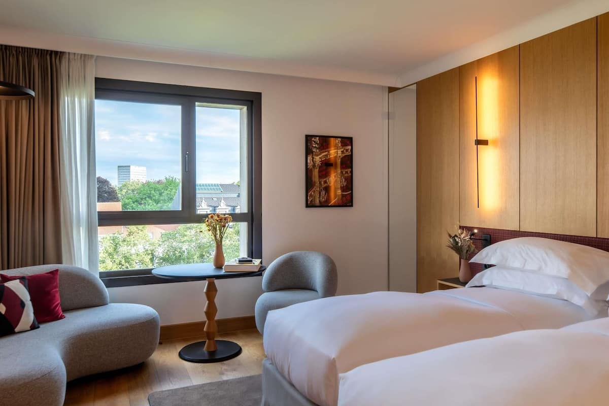 Una habitación de hotel con cama, sofá, sillón, una mesita con un florero y ventana con vistas a edificios y árboles.