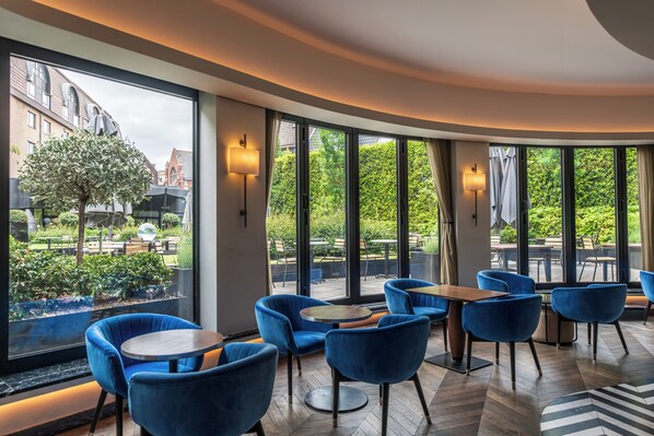 Un espace de détente intérieur moderne, doté de grandes fenêtres, de chaises bleues et d’un parquet disposé en motif chevrons.
