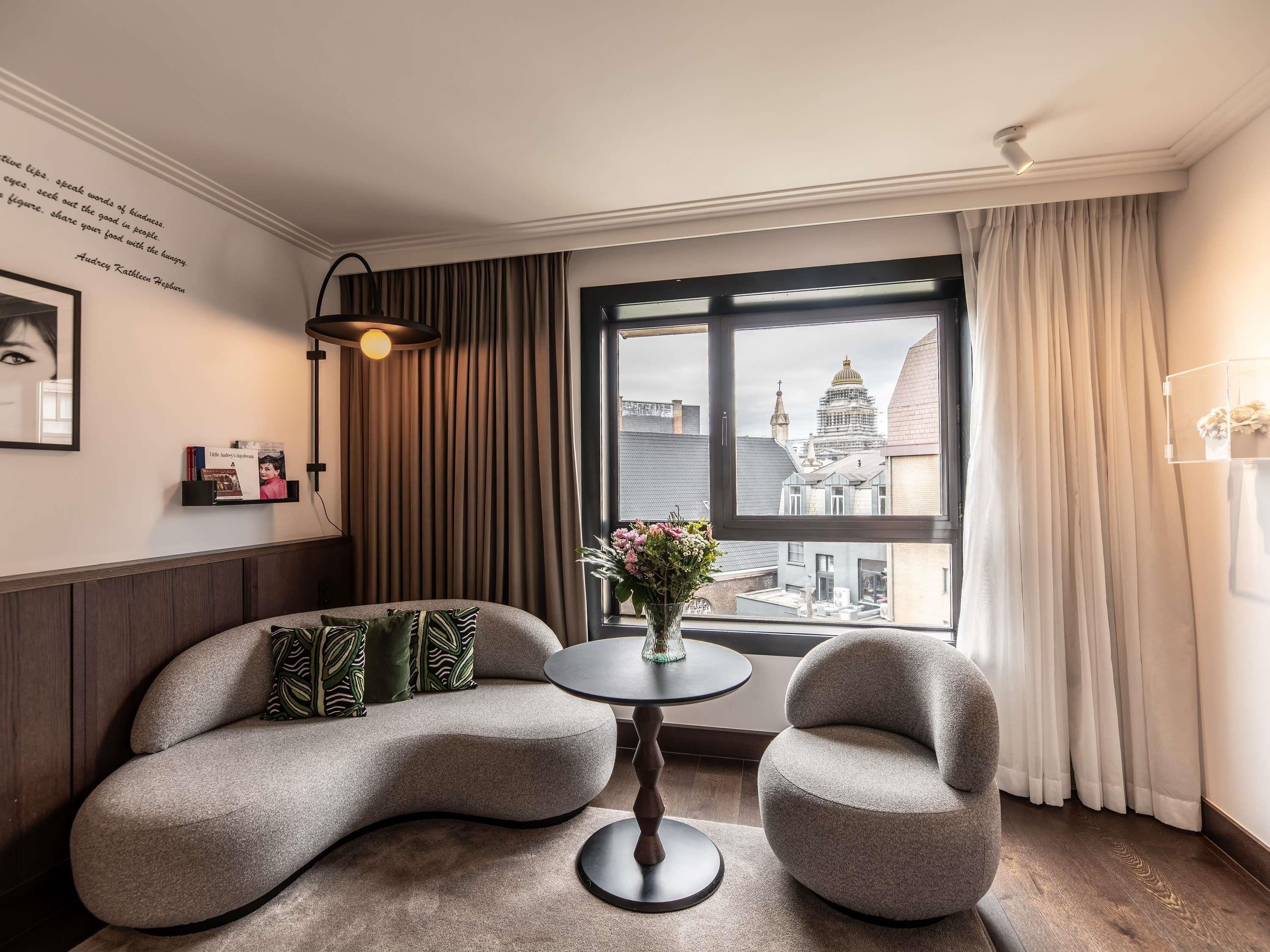 Ein modernes Zimmer mit einer gebogenen Couch, zwei Sesseln, einem kleinen runden Tisch und Blick auf eine Skyline durch ein großes Fenster.