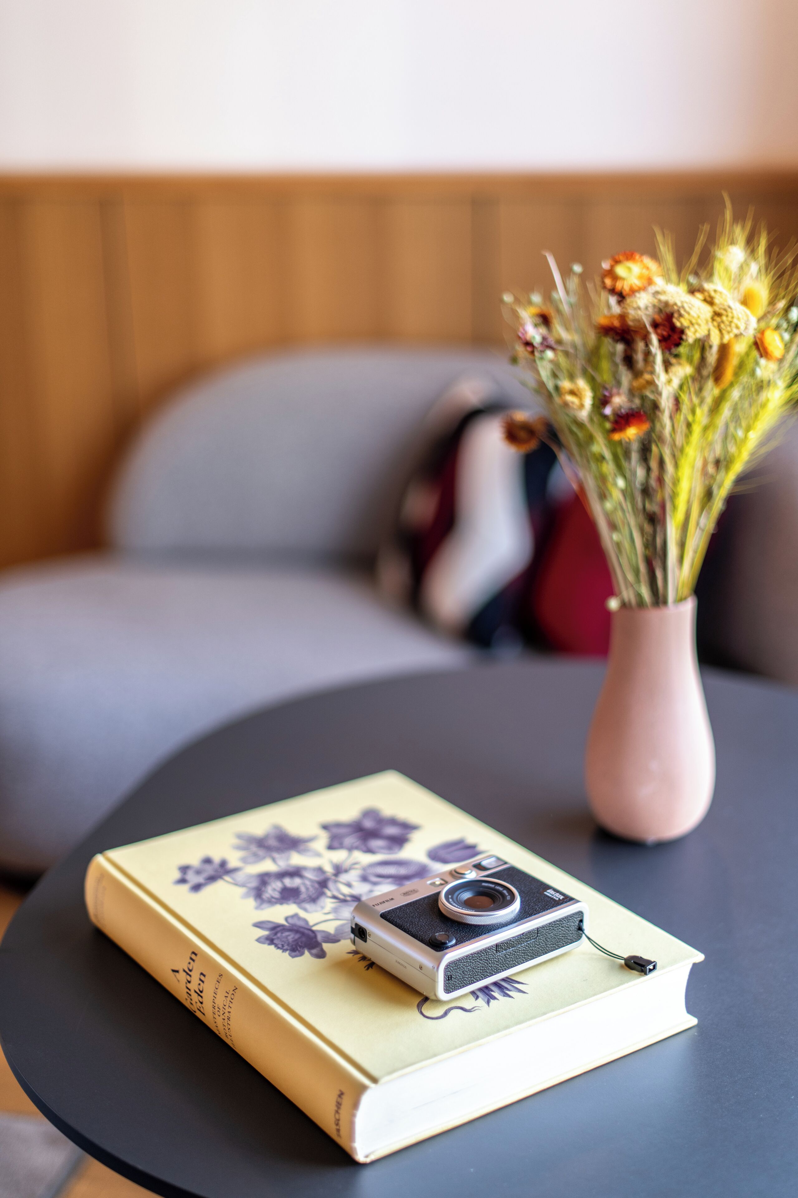 Ein Buch mit einer Kamera darauf, eine Vase mit Blumen und eine Couch im Hintergrund.