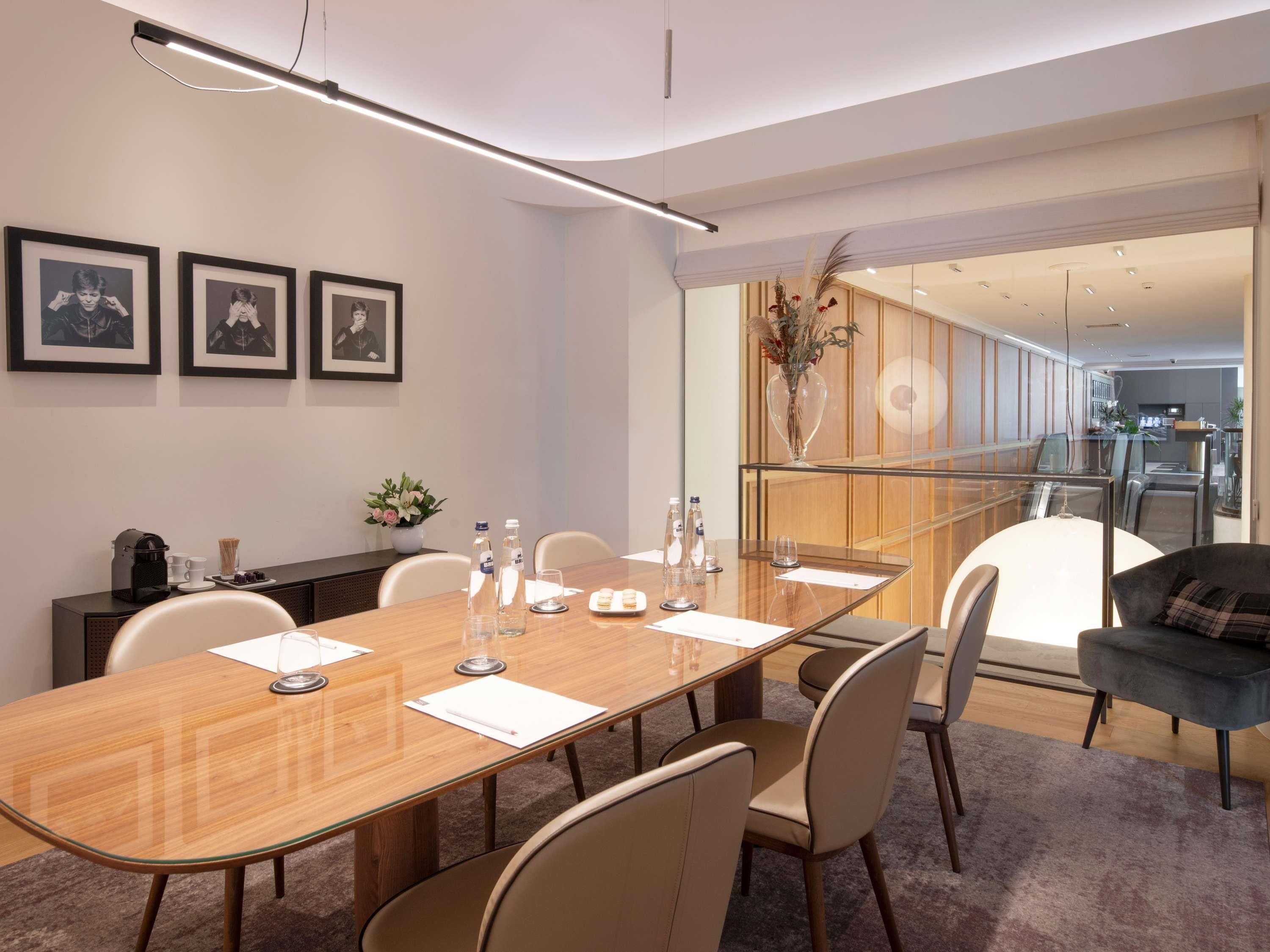 A conference room with a long table, chairs, framed pictures on the wall, a vase with flowers, and a glass partition leading to a kitchen area.