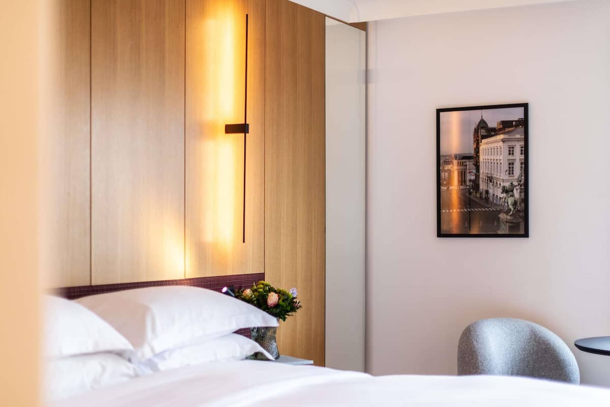 A hotel room with a bed, a nightstand with a vase of flowers, a framed cityscape picture on the wall, and a chair.