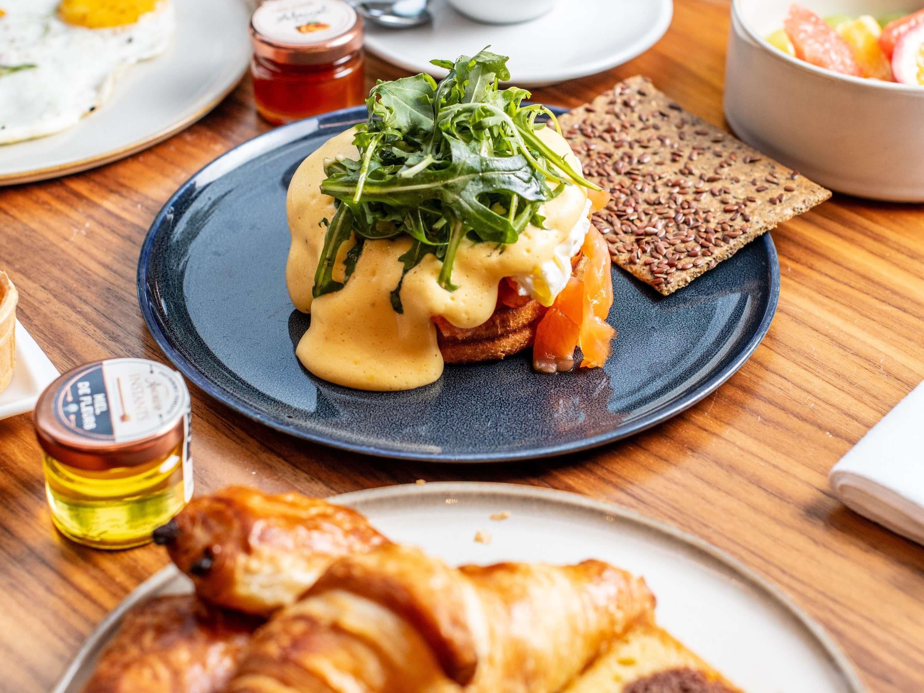 A breakfast setting with a plate of eggs benedict, a croissant, and a bowl of fruit salad.