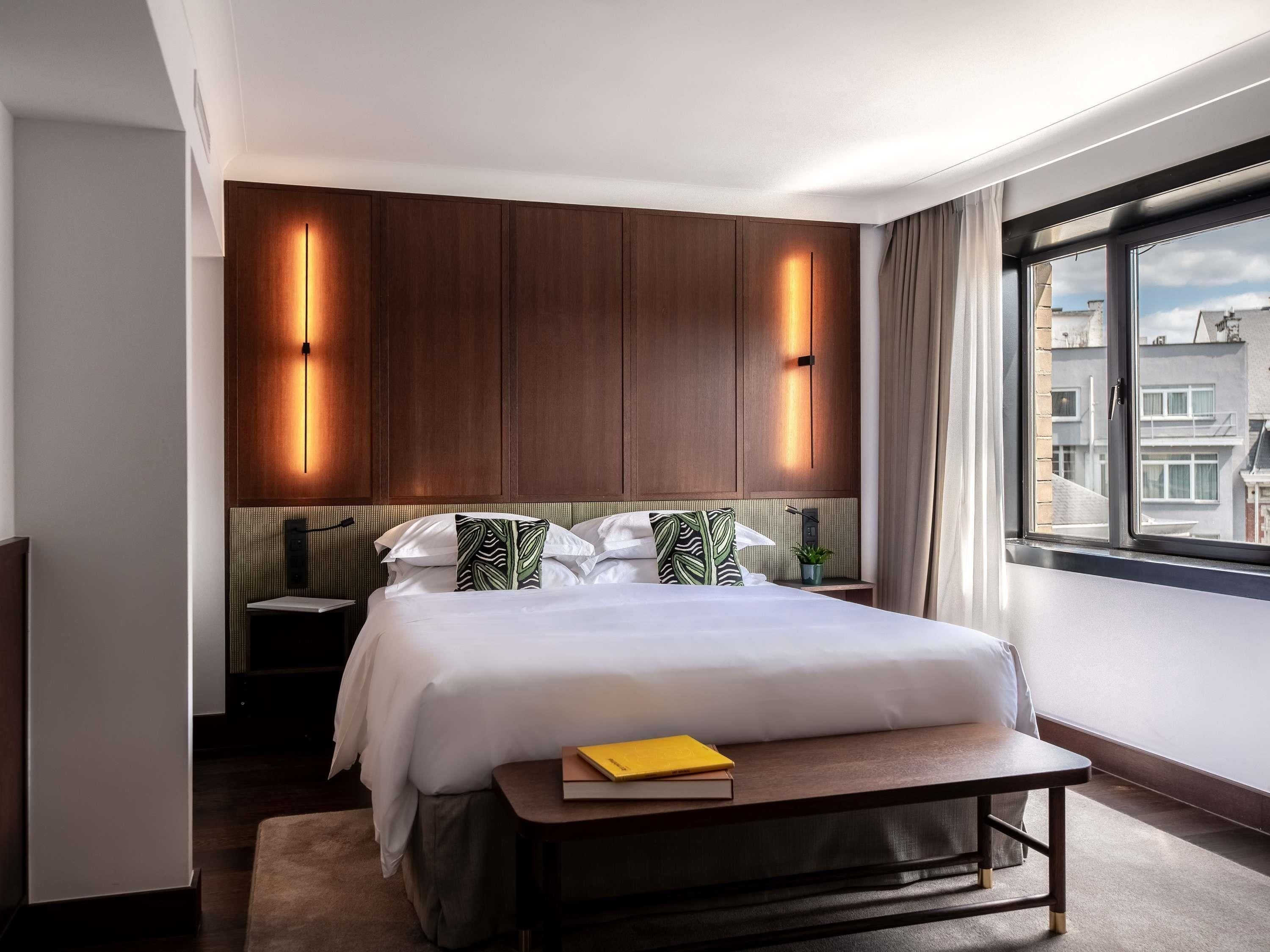 Une chambre d’hôtel moderne avec un grand lit, des tables de chevet, une lampe de chevet, une fenêtre donnant sur des immeubles et un luminaire fixé au mur.
