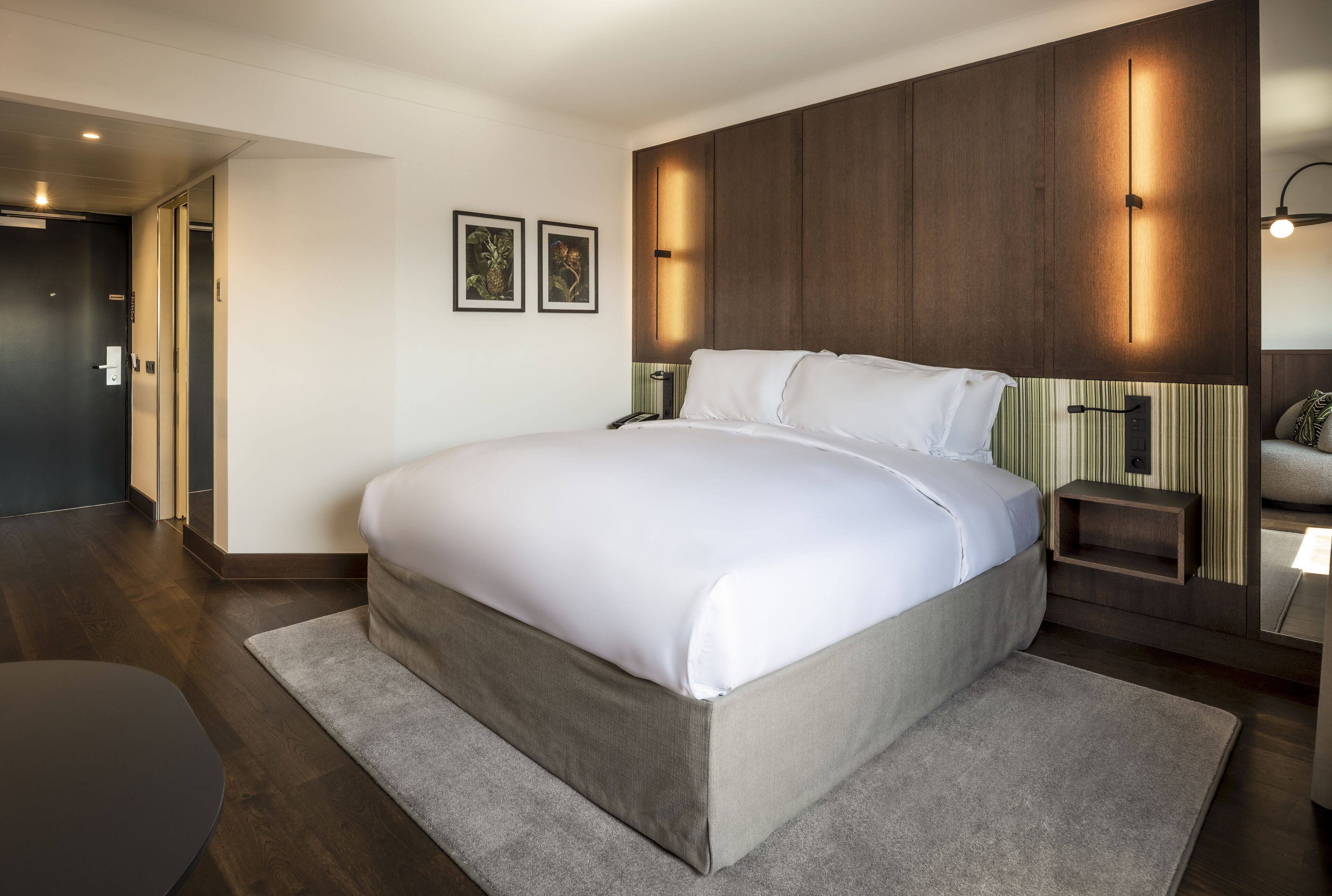 A modern hotel room with a large bed, a bedside table, a nightstand, and a wall-mounted light fixture.