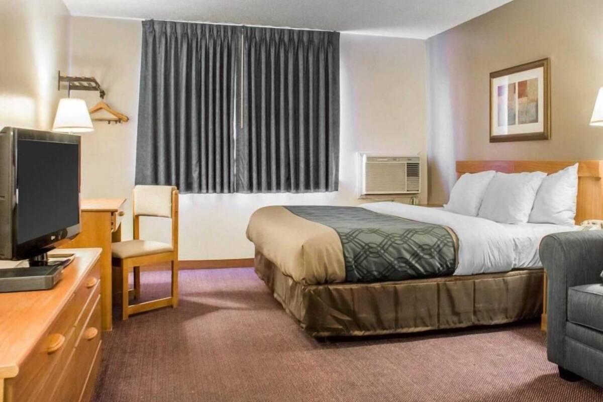 A hotel room with a bed, a desk with a television, a chair, a bedside table, a lamp, an air conditioner, and a framed picture on the wall.
