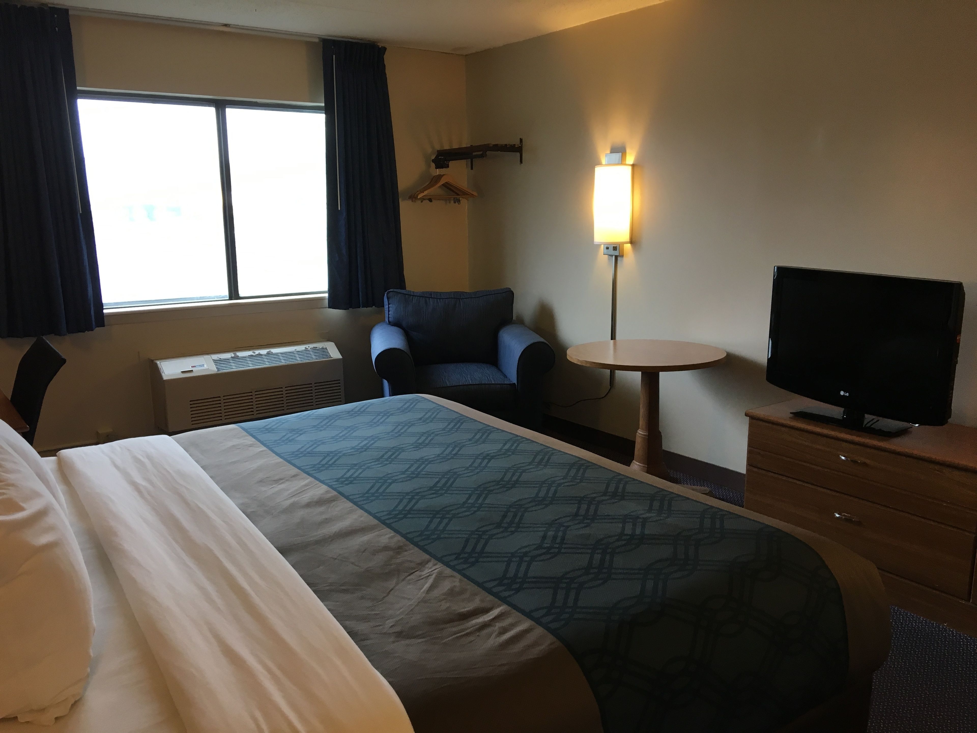 Una habitación de hotel con cama, una silla, televisión, una mesita y una lámpara fijada a la pared.