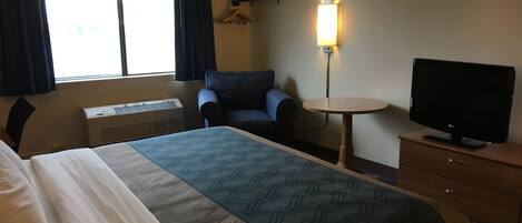 Una habitación de hotel con cama, una silla, televisión, una mesita y una lámpara fijada a la pared.