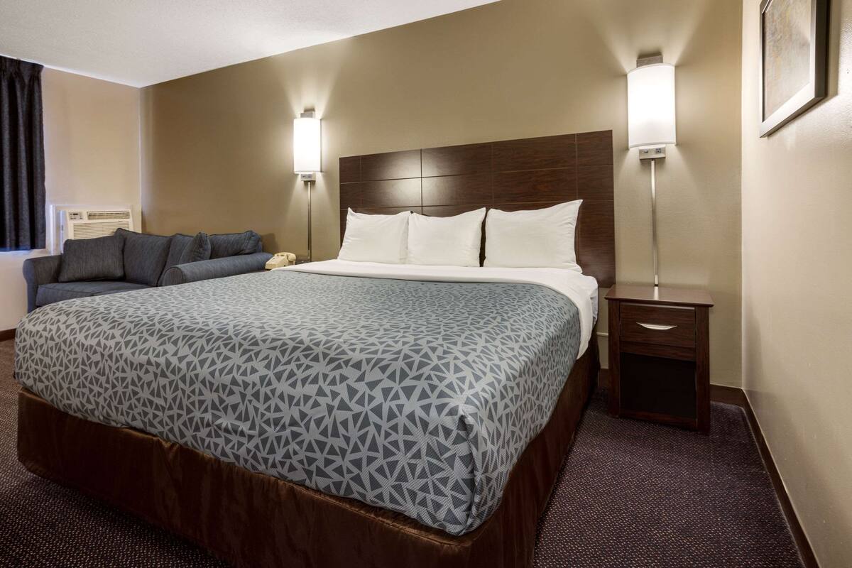 A hotel room with a large bed, a sofa, two bedside lamps, a nightstand, and a window with curtains.